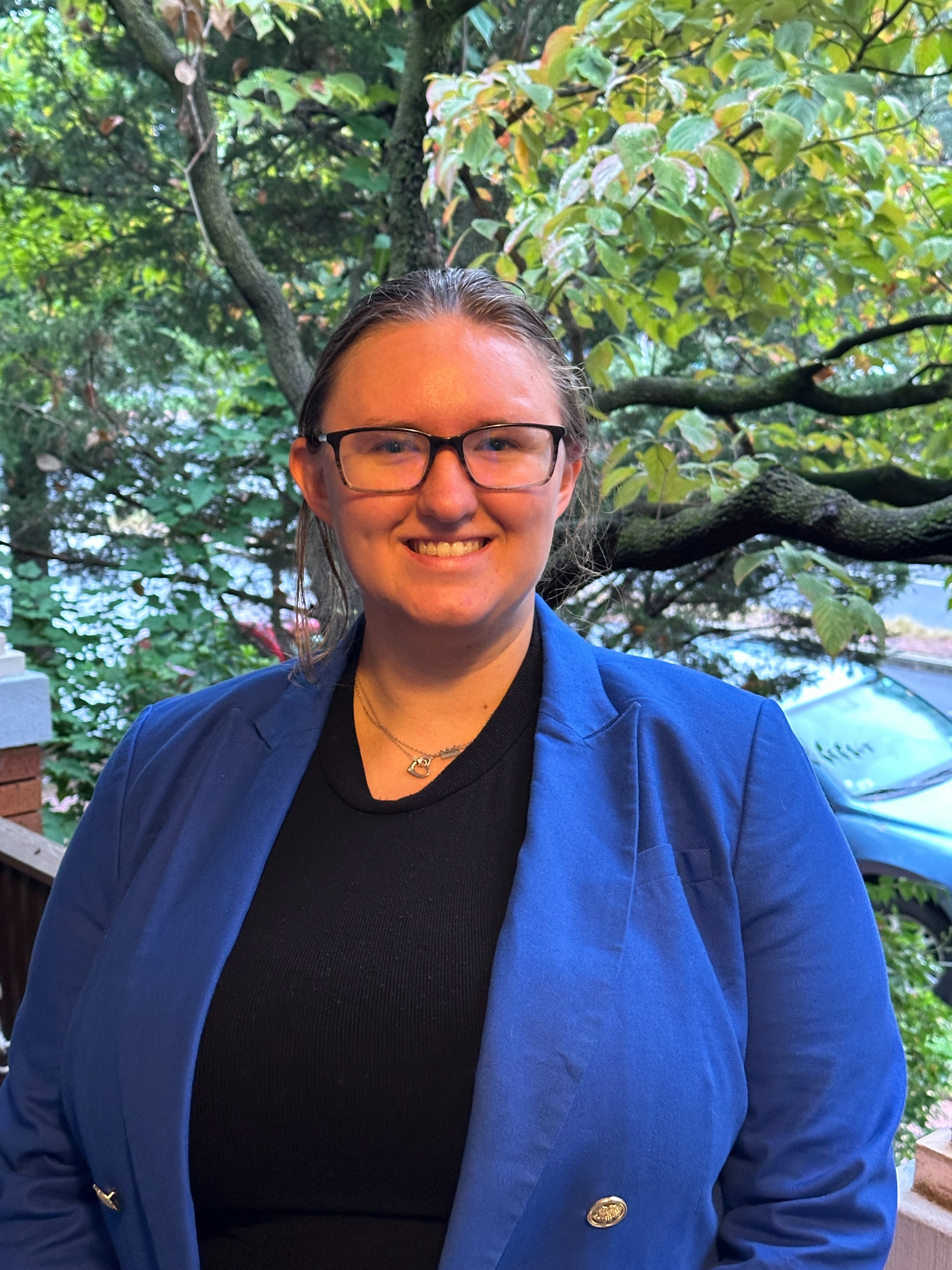 Woman stands in front of tree wearing black shirt, blue blazer, and glasses.